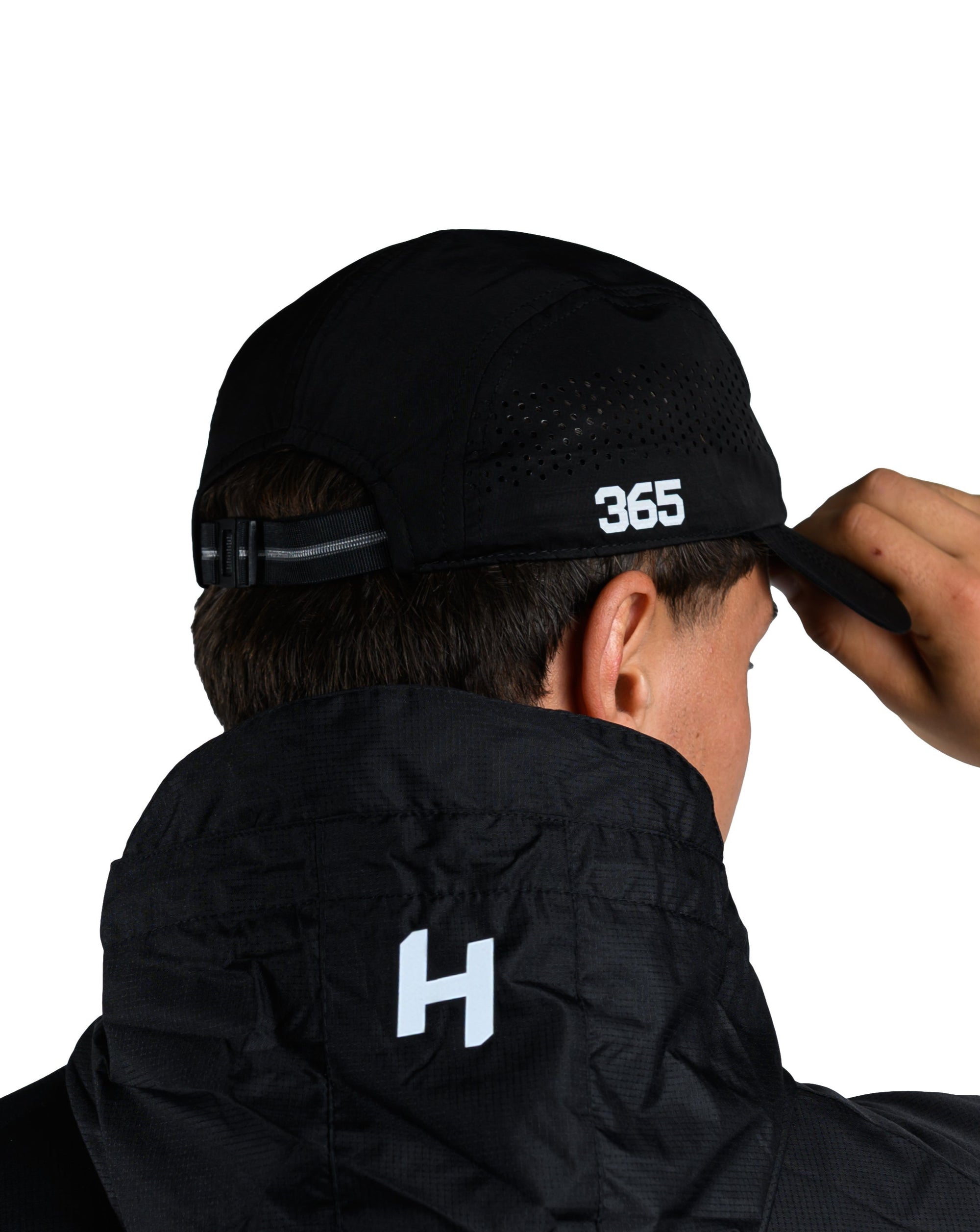 Person wearing a black jacket with '365' printed on the back, adjusting a black cap.