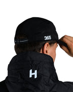 Person wearing a black jacket with '365' printed on the back, adjusting a black cap.